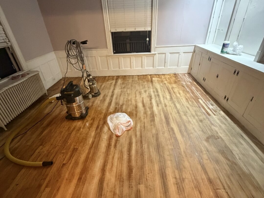 The same room mid-project from the opposite corner: floor sanders on stripped boards and lilac walls above the wainscoting